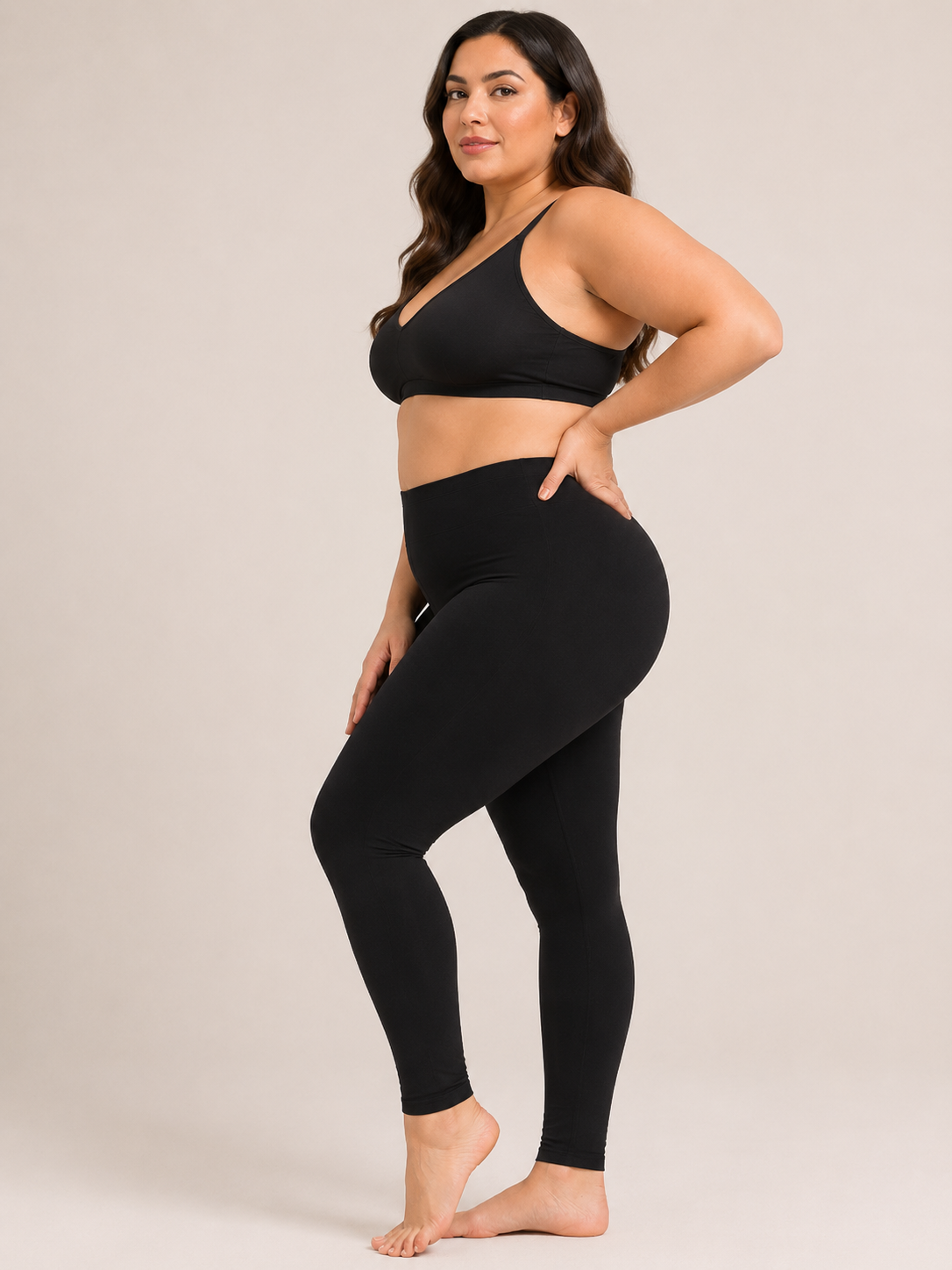 A woman modeling a matching black activewear set consisting of a sports bra and high-waisted leggings, standing against a solid light beige studio background.