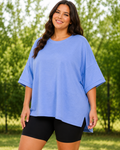 A woman wearing an oversized light blue short-sleeve t-shirt and black bike shorts, standing outdoors in front of a blurred green tree background.