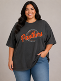 A woman wearing a dark charcoal gray graphic t-shirt with 'Panthers' written in orange script inside a distressed circular logo, paired with blue jeans, against a solid gray studio background.
