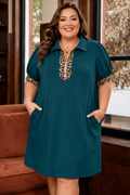 A woman modeling a teal short-sleeve collared dress with leopard print trim at the neckline and sleeve cuffs, standing indoors near a window and brown sofa.