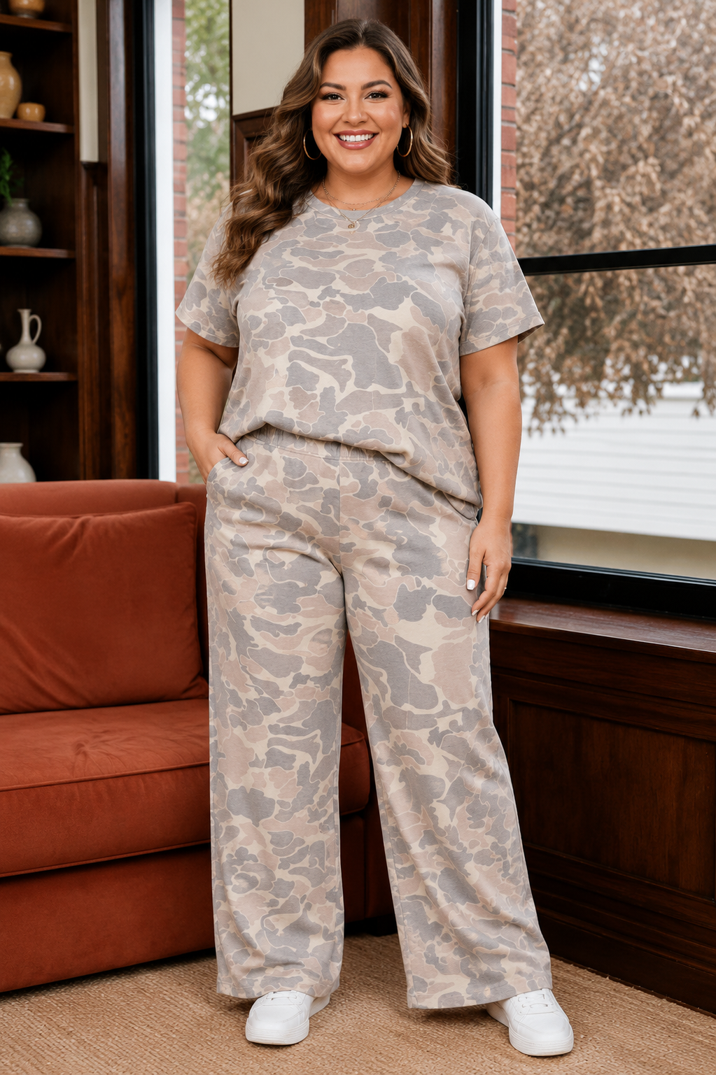 A woman modeling a matching two-piece lounge set with a beige and gray camouflage pattern, standing in a cozy room with a rust-colored sofa and a window.