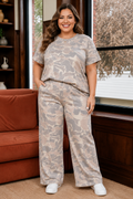 A woman modeling a matching two-piece lounge set with a beige and gray camouflage pattern, standing in a cozy room with a rust-colored sofa and a window.