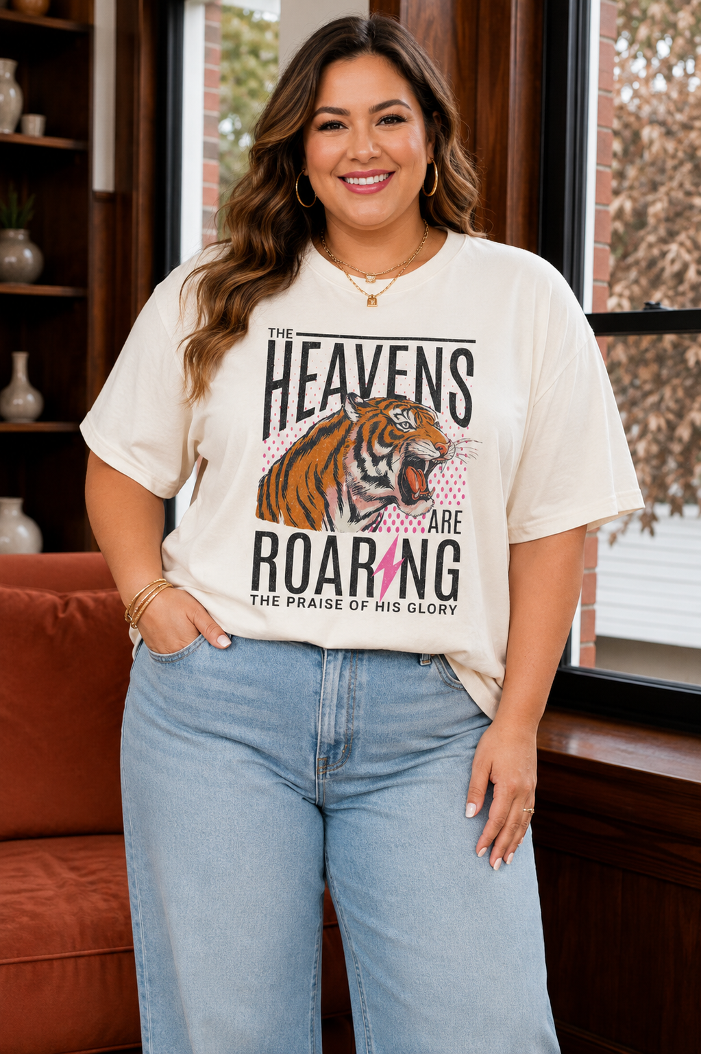 A woman wearing a cream-colored graphic t-shirt with a tiger illustration and the text 'THE HEAVENS ARE ROARING' paired with light blue jeans, standing in a cozy indoor setting.