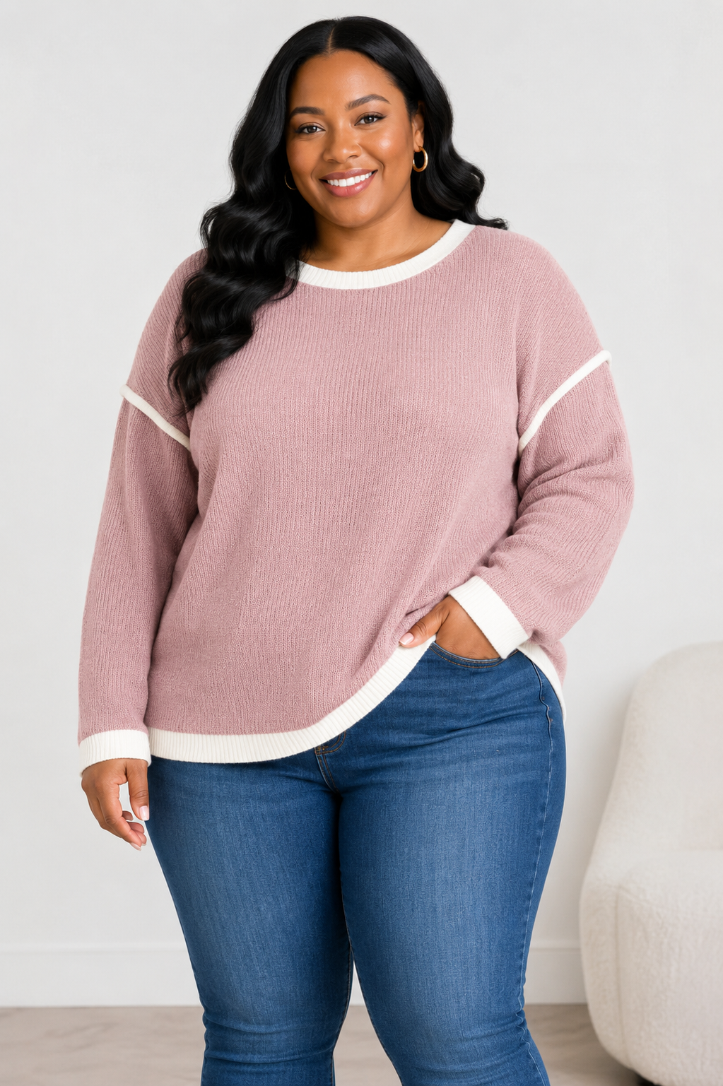 A woman wearing a dusty rose ribbed knit sweater with white contrast trim on the collar, cuffs, and hem, paired with blue jeans, standing in a brightly lit room.
