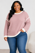 A woman wearing a dusty rose ribbed knit sweater with white contrast trim on the collar, cuffs, and hem, paired with blue jeans, standing in a brightly lit room.