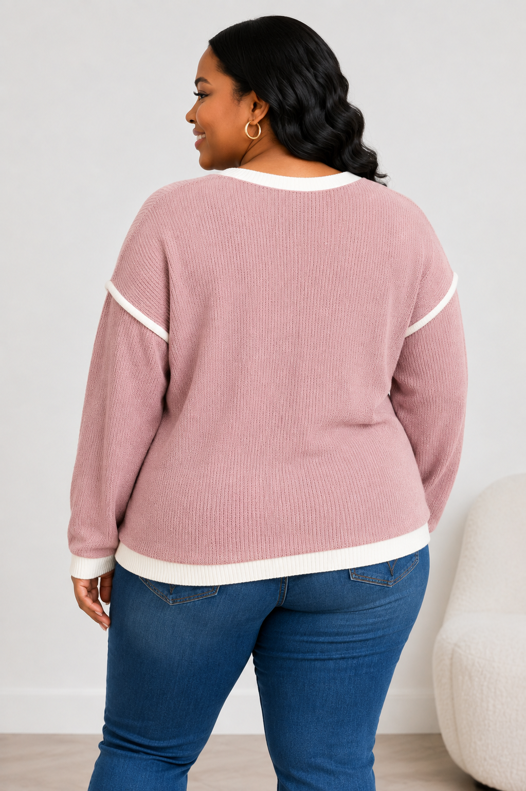 A woman shown from the back wearing a dusty pink ribbed knit sweater with white contrast trim, paired with blue jeans, in a brightly lit indoor setting.