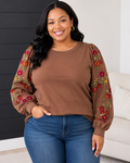 A woman wearing a brown crewneck sweatshirt with long sheer sleeves decorated with red, pink, and yellow floral embroidery, paired with blue jeans.