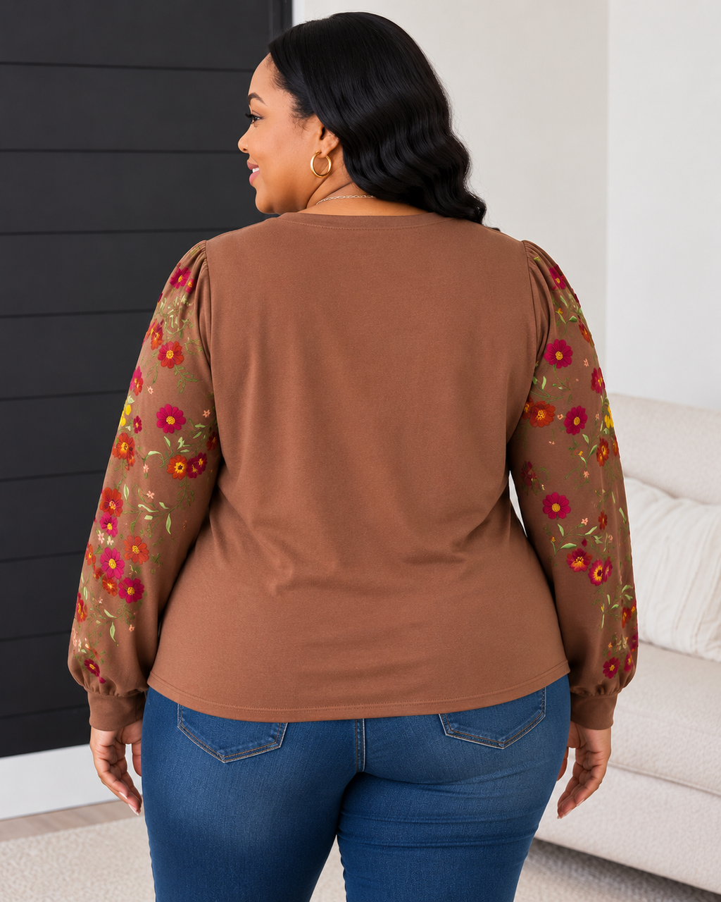 A woman shown from the back wearing a brown long-sleeve top with sheer floral-patterned sleeves and blue jeans, standing indoors near a sofa and dark paneled wall.