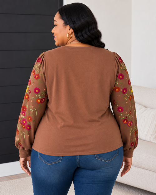 A woman shown from the back wearing a brown long-sleeve top with sheer floral-patterned sleeves and blue jeans, standing indoors near a sofa and dark paneled wall.