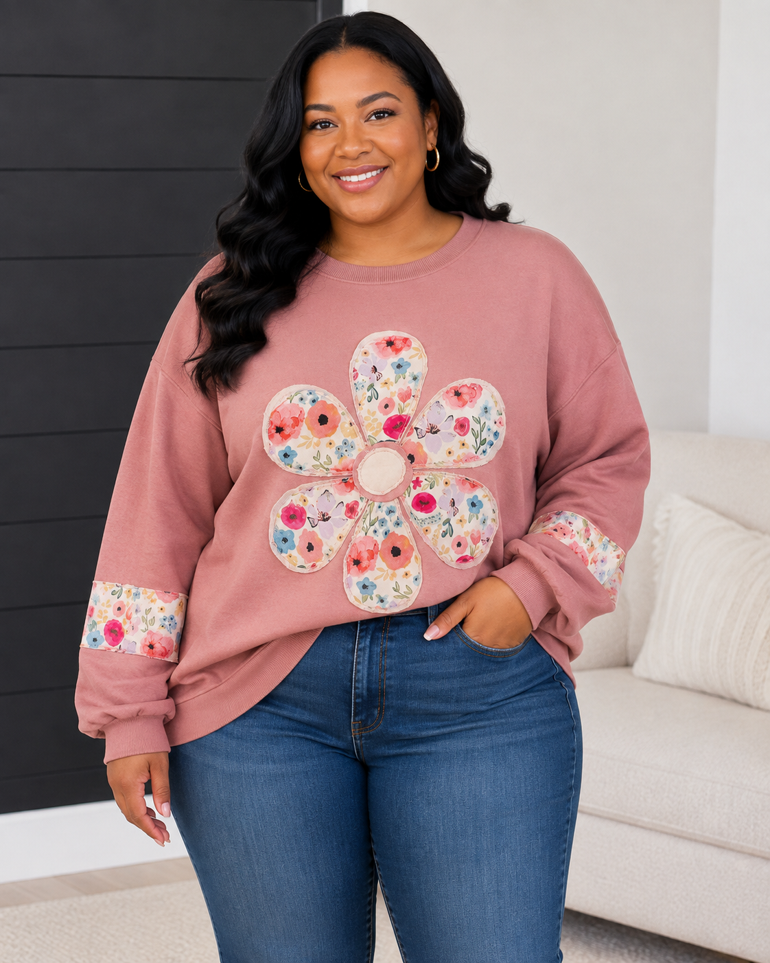 A woman wearing a dusty rose crewneck sweatshirt with a large floral applique on the front and matching floral trim on the cuffs, paired with blue jeans in a modern indoor setting.