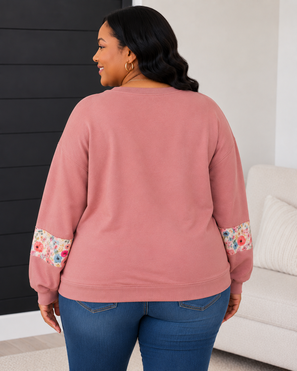 A woman shown from the back wearing a dusty rose crewneck sweatshirt with colorful floral patterned cuffs, paired with blue jeans in a modern indoor setting.