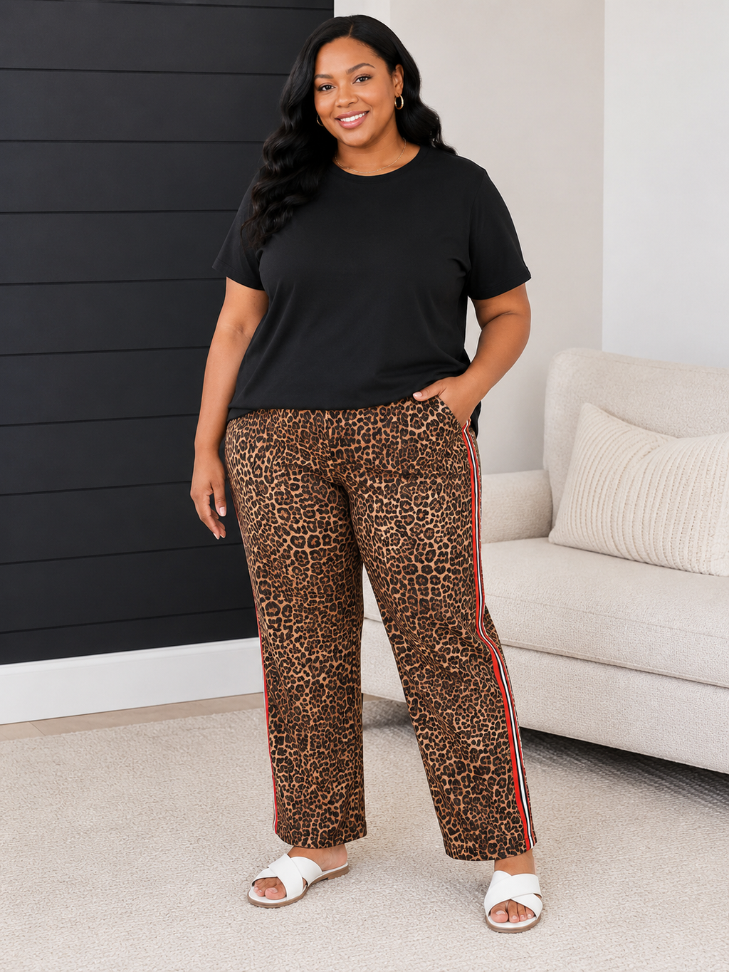 A woman modeling leopard-print pants with red and blue side stripes, paired with a black t-shirt and white slide sandals, standing in a living room setting.