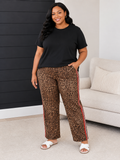 A woman modeling leopard-print pants with red and blue side stripes, paired with a black t-shirt and white slide sandals, standing in a living room setting.
