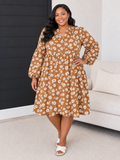 A woman modeling a brown long-sleeve midi dress with a white daisy floral pattern, standing in a living room setting next to a light-colored sofa.