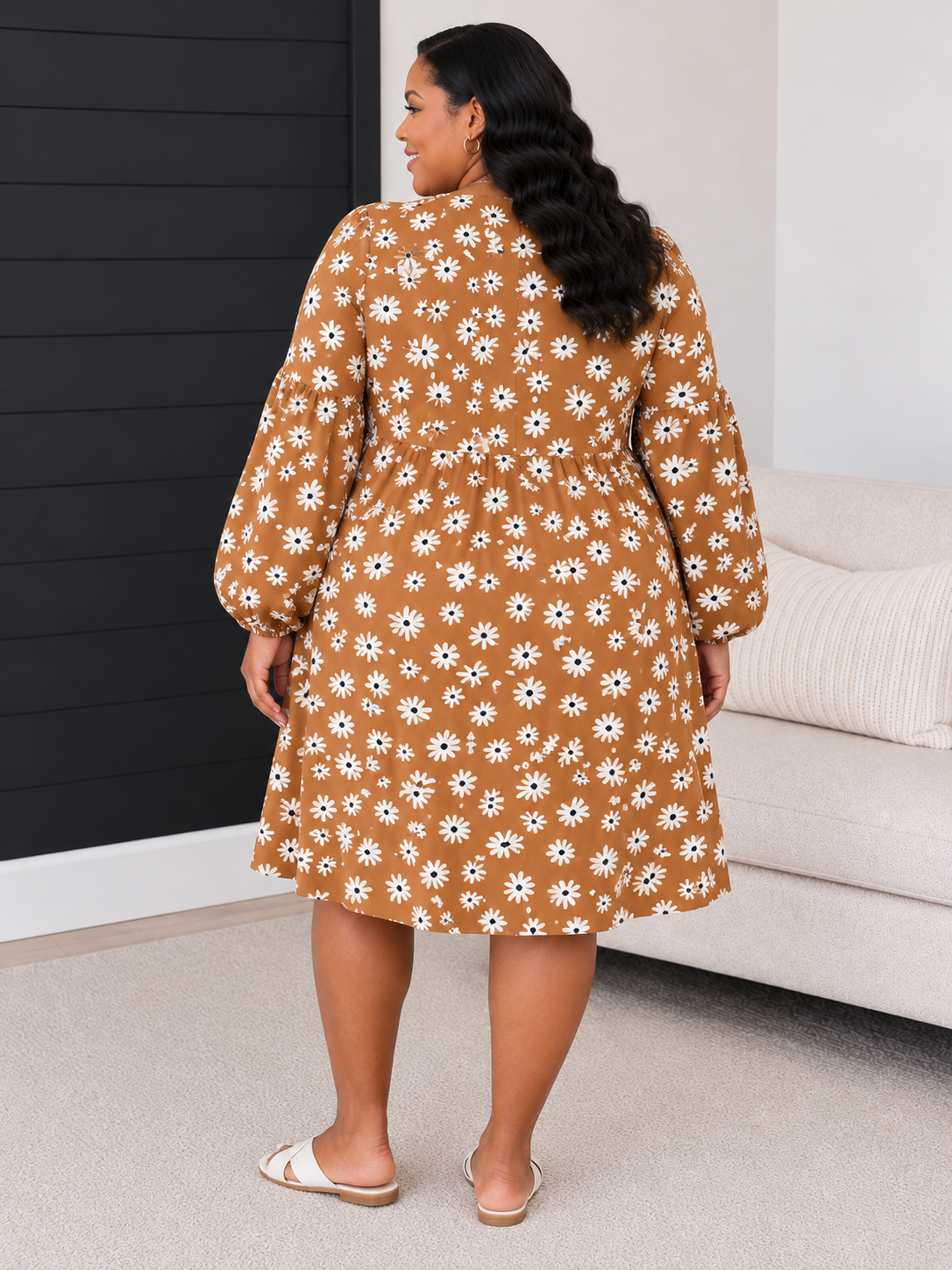 A woman wearing a brown long-sleeve midi dress with a white daisy floral pattern, shown from the back in a living room setting.