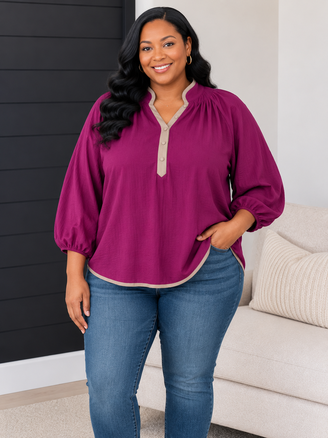 A woman modeling a magenta long-sleeve blouse with a beige trim and button placket, paired with blue jeans in a modern living room setting.
