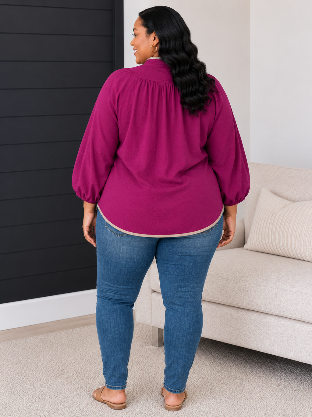 A woman wearing a magenta long-sleeve blouse and blue jeans, shown from the back in a living room setting with a sofa and dark paneled wall.