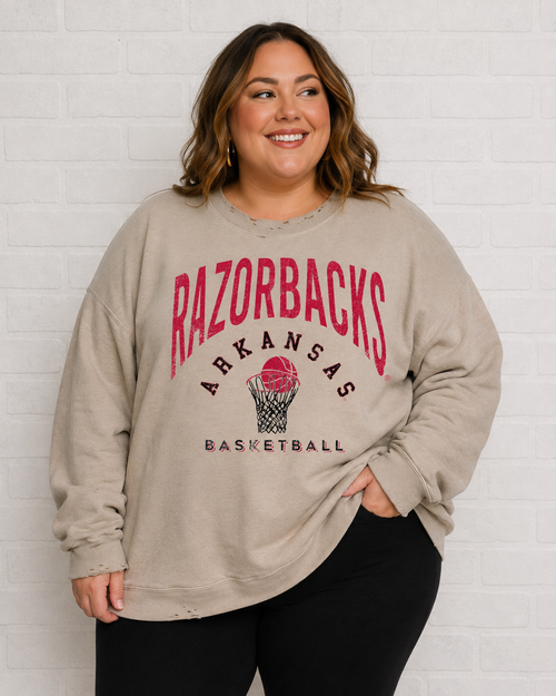 A woman wearing a beige Arkansas Razorbacks basketball sweatshirt with a red and black graphic print, paired with black pants, standing against a white brick wall.