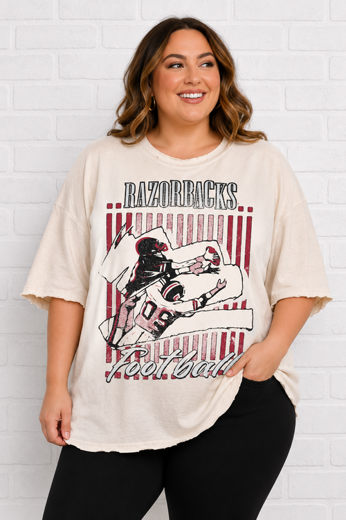A woman modeling a cream-colored, oversized t-shirt with a distressed graphic that reads 'RAZORBACKS Football' and features a football player illustration, set against a white brick wall background.