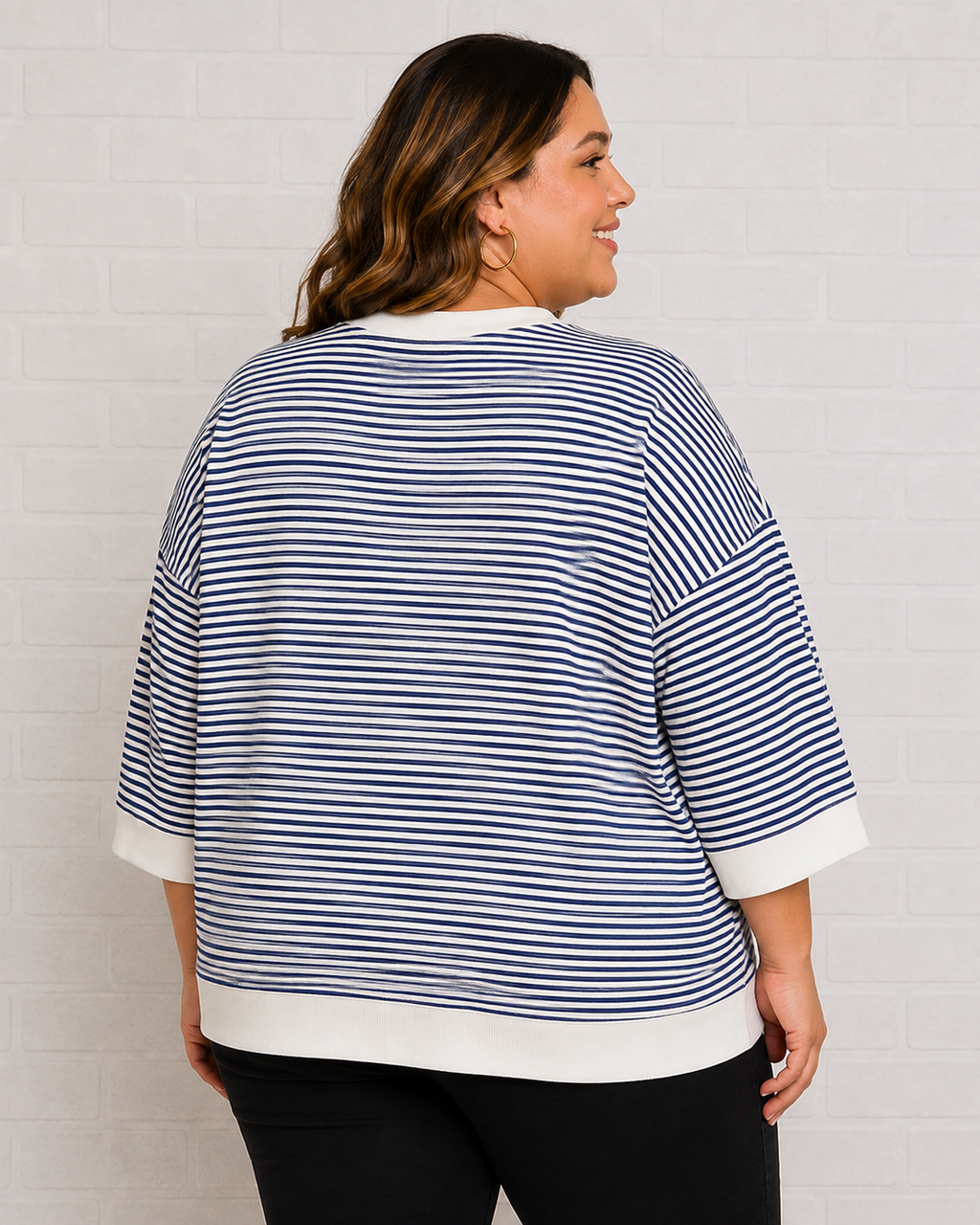 A woman shown from the back wearing a navy and white horizontally striped knit top with white ribbed trim, paired with black pants, standing against a white brick wall.