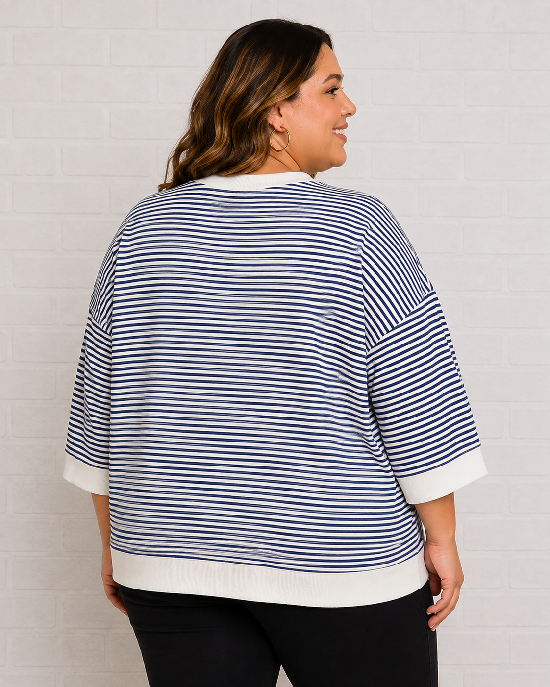 A woman shown from the back wearing a navy and white horizontally striped knit top with white ribbed trim, paired with black pants, standing against a white brick wall.