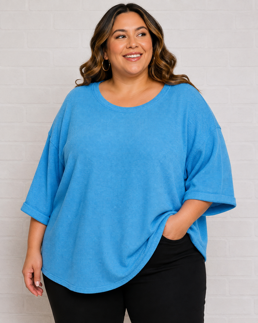 A woman wearing a bright blue oversized short-sleeve top with three-quarter sleeves and black pants, standing against a white brick wall.