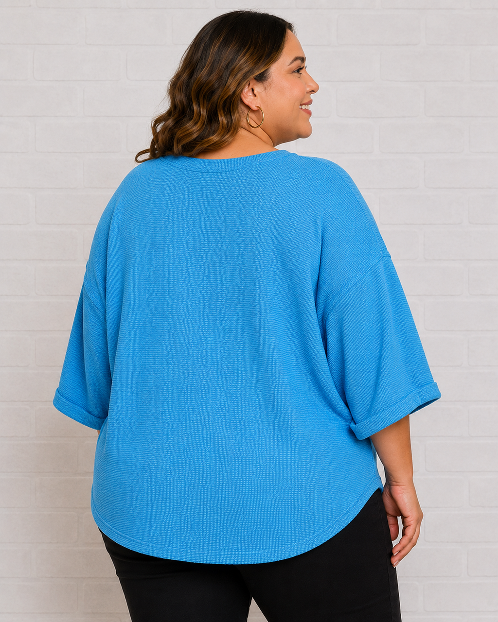 A woman shown from the back wearing a bright blue oversized short-sleeve top and black pants, standing against a white brick wall.