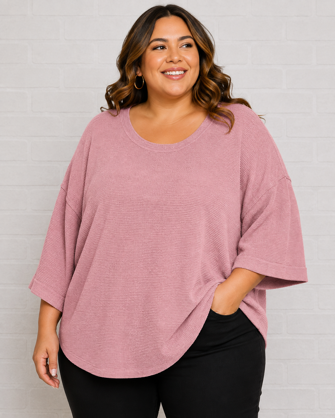 A woman wearing a dusty rose pink oversized short-sleeve top with black pants, standing against a white brick wall.