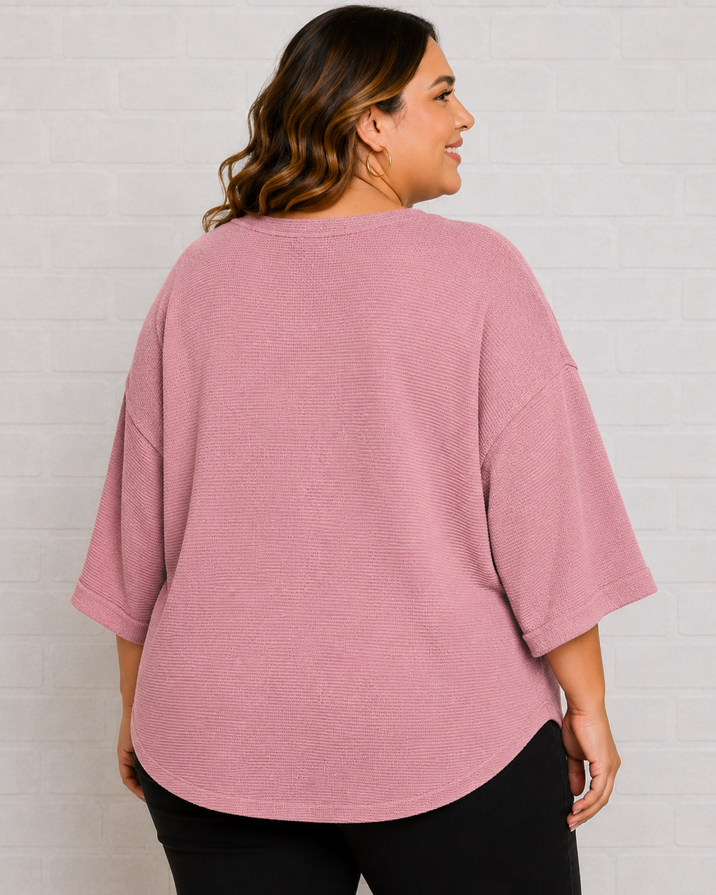 A woman shown from the back wearing a dusty pink waffle-knit short-sleeve top and black pants, standing against a white brick wall.
