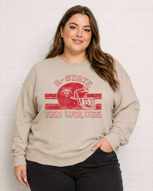 A woman wearing a beige crewneck sweatshirt with a red distressed graphic that reads 'A-STATE RED WOLVES' and shows a football helmet illustration, standing against a white brick wall.
