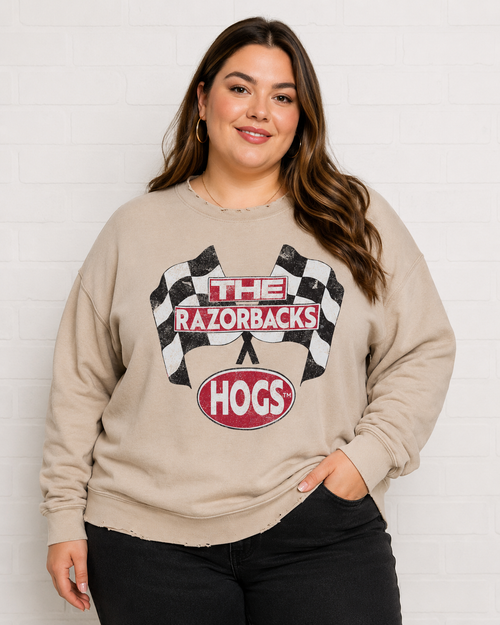 A woman wearing a beige crewneck sweatshirt with a 'The Razorbacks Hogs' racing graphic, paired with black pants, standing against a white brick wall.