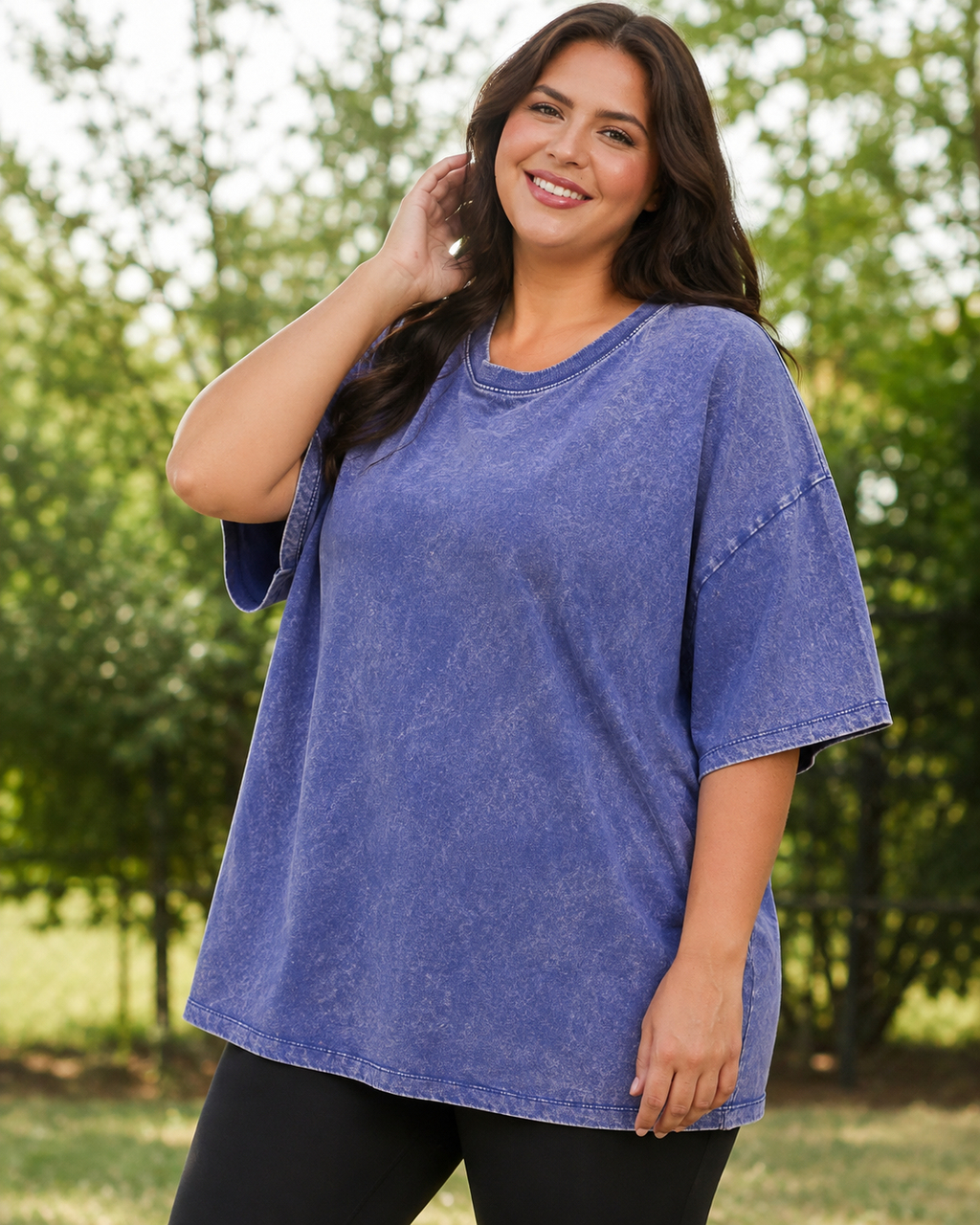 A woman wearing a short-sleeve, oversized crewneck t-shirt in a washed periwinkle blue color, standing outdoors in front of a blurred green background.