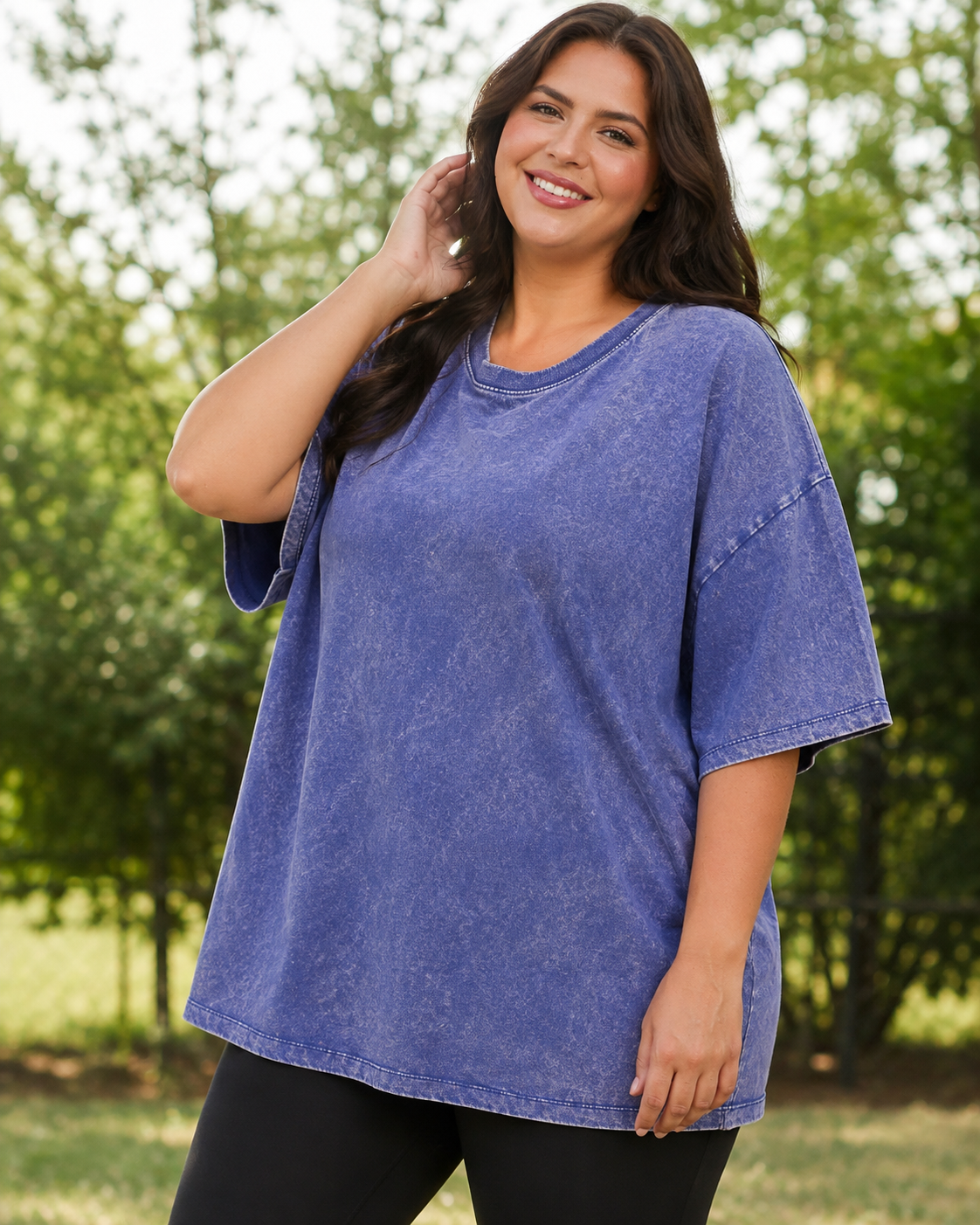 A woman wearing a short-sleeve, oversized crewneck t-shirt in a washed periwinkle blue color, standing outdoors in front of a blurred green background.