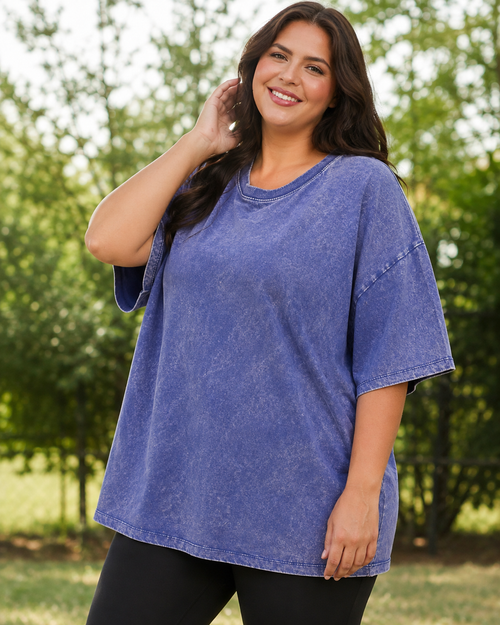 A woman wearing a short-sleeve, oversized crewneck t-shirt in a washed periwinkle blue color, standing outdoors in front of a blurred green background.