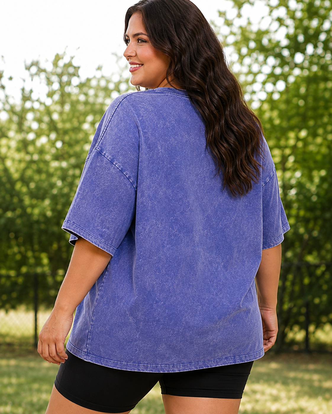 A woman wearing an oversized washed blue t-shirt and black shorts, photographed from behind in an outdoor park setting with green trees in the background.