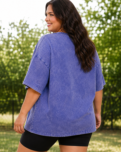 A woman wearing an oversized washed blue t-shirt and black shorts, photographed from behind in an outdoor park setting with green trees in the background.