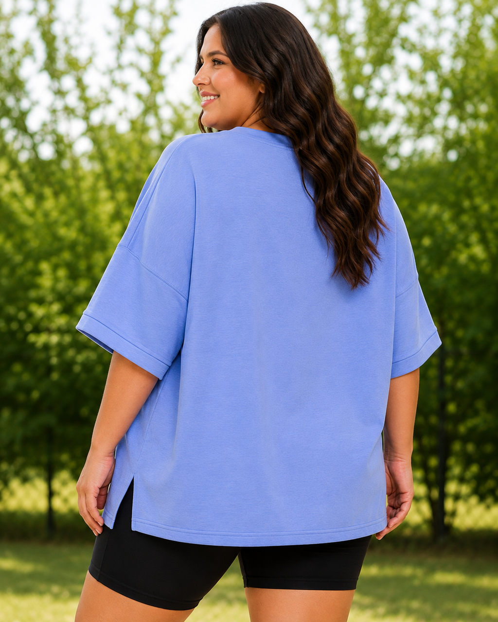 A woman wearing an oversized light blue t-shirt and black bike shorts, photographed from behind in an outdoor park setting with green trees in the background.