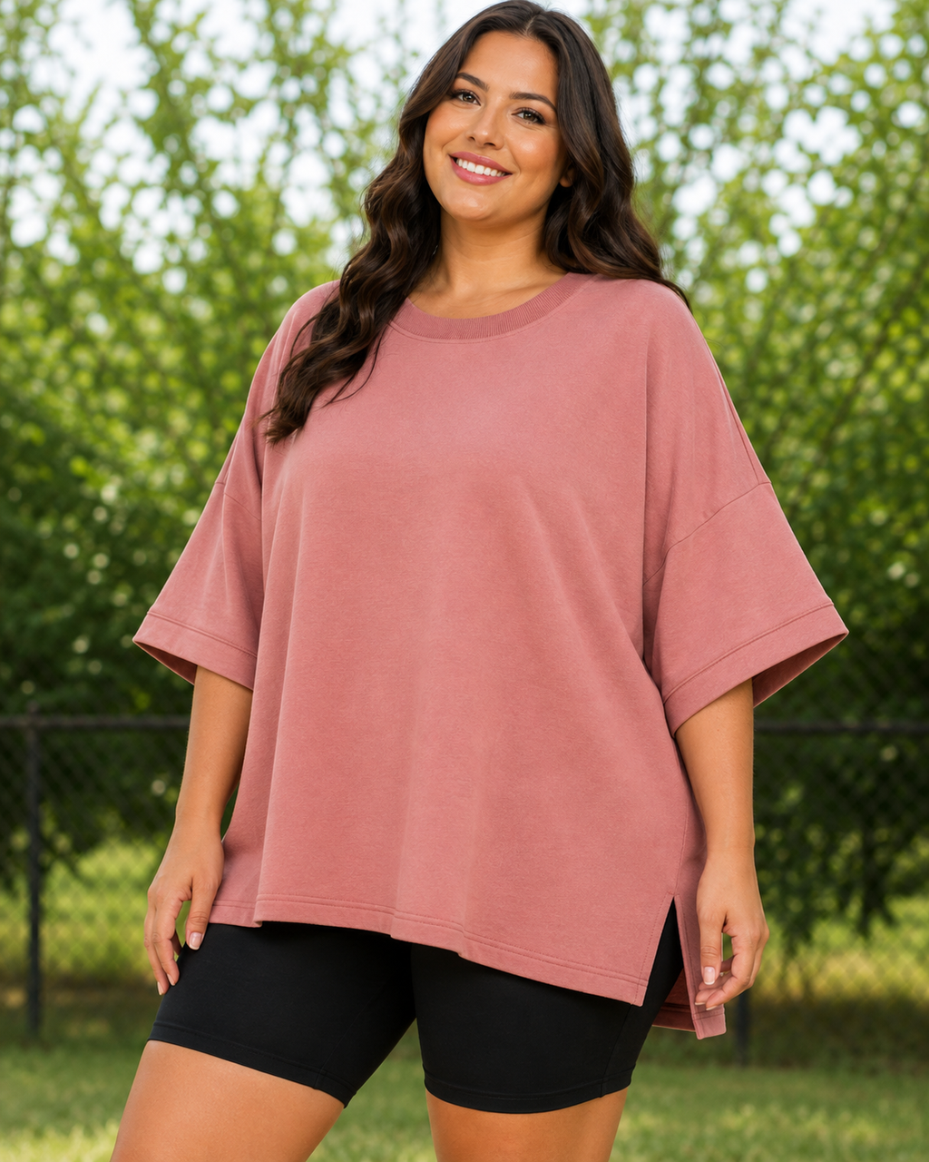 A woman wearing an oversized dusty rose short-sleeve t-shirt and black bike shorts, standing outdoors in front of a green tree background.