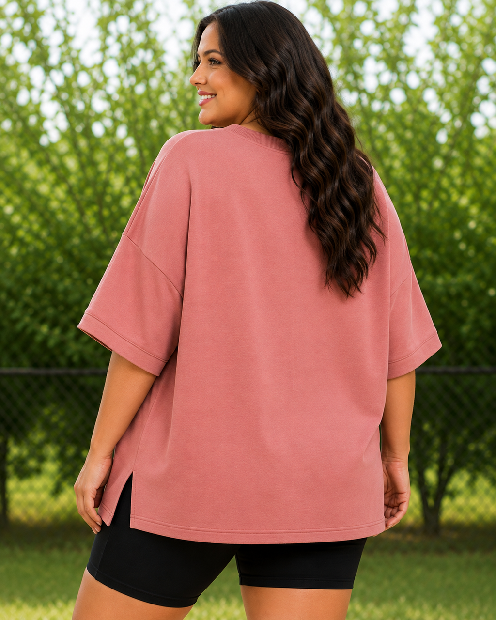A woman wearing an oversized dusty rose short-sleeve t-shirt and black bike shorts, photographed from behind in an outdoor park setting with green trees in the background.