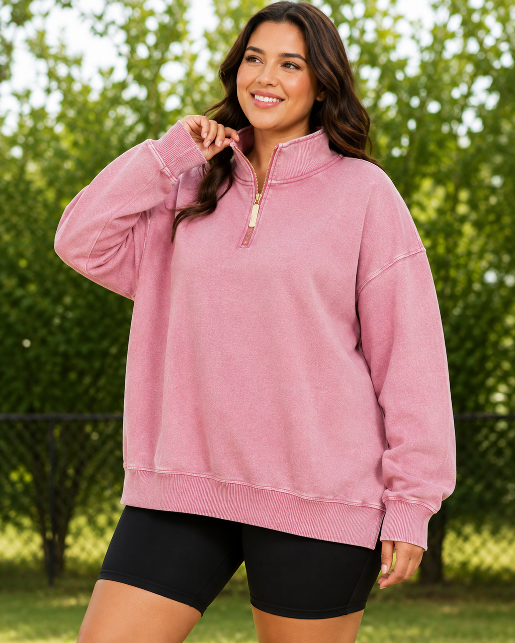 A woman wearing an oversized pink quarter-zip sweatshirt and black bike shorts, standing outdoors in front of a blurred green hedge.