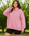 A woman wearing an oversized pink quarter-zip sweatshirt and black bike shorts, standing outdoors in front of a blurred green hedge.