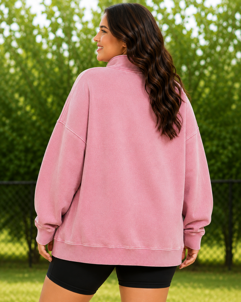 A woman wearing an oversized dusty pink sweatshirt, shown from the back in an outdoor setting with green trees in the background.