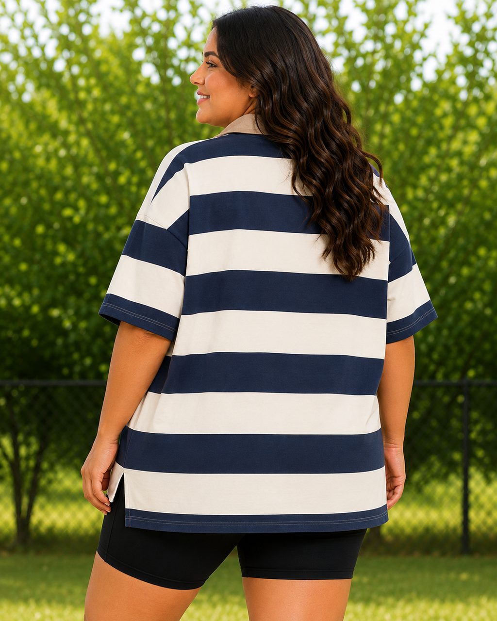 A woman wearing an oversized short-sleeve polo shirt with navy blue and off-white horizontal stripes, photographed outdoors with green trees in the background.