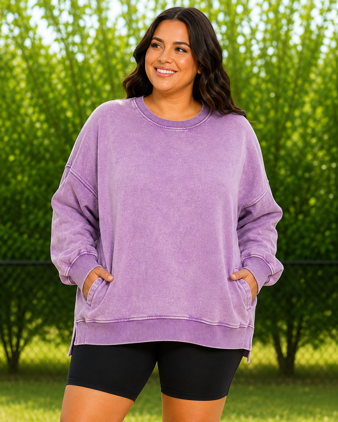 A woman wearing an oversized lavender-purple washed sweatshirt with side slits and front pockets, paired with black bike shorts, standing outdoors in front of a green hedge.