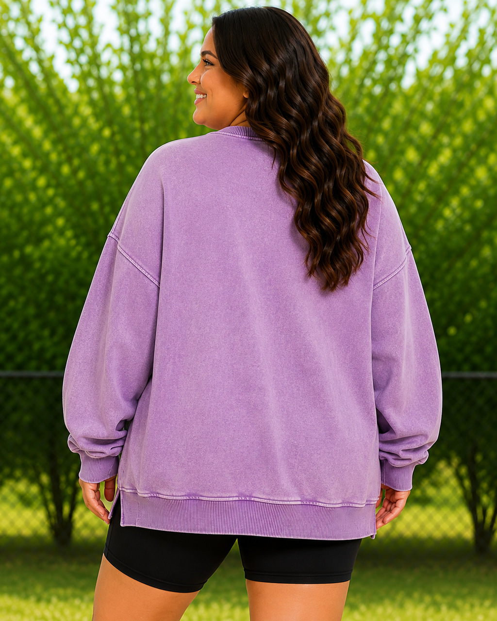 A woman viewed from behind wearing an oversized lavender-purple sweatshirt and black shorts, standing outdoors in front of green trees.