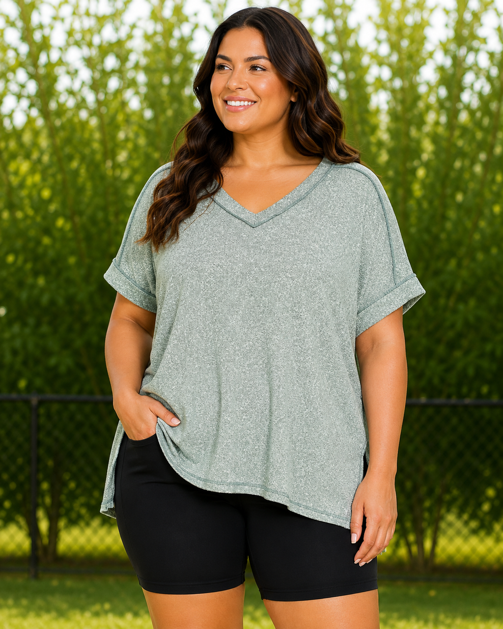 A woman wearing a heathered sage green short-sleeve V-neck athletic top and black bike shorts, standing outdoors in front of a green hedge and chain-link fence.