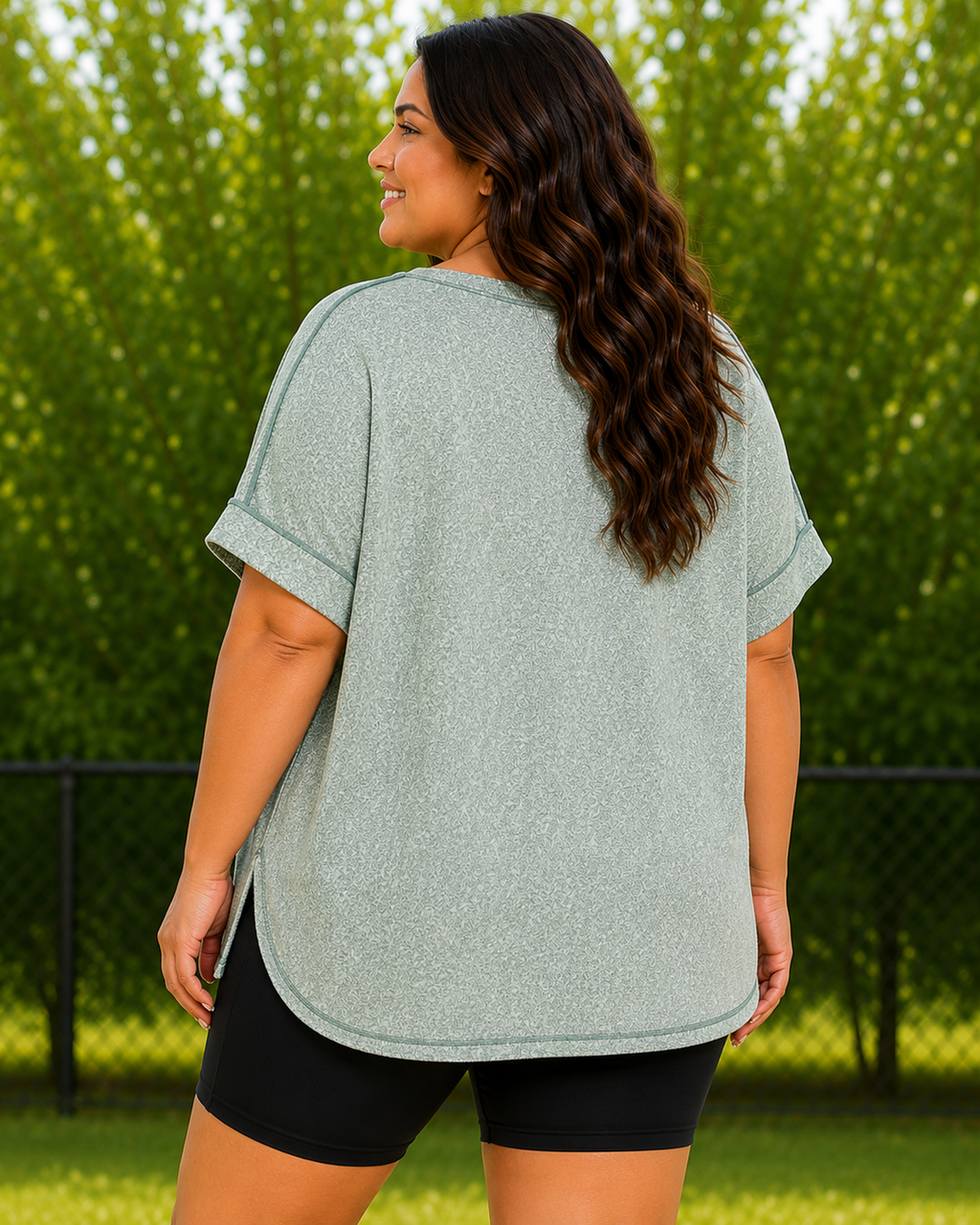 A woman wearing a heathered sage green short-sleeve top and black athletic shorts, photographed from the back in an outdoor park setting with green trees and grass.