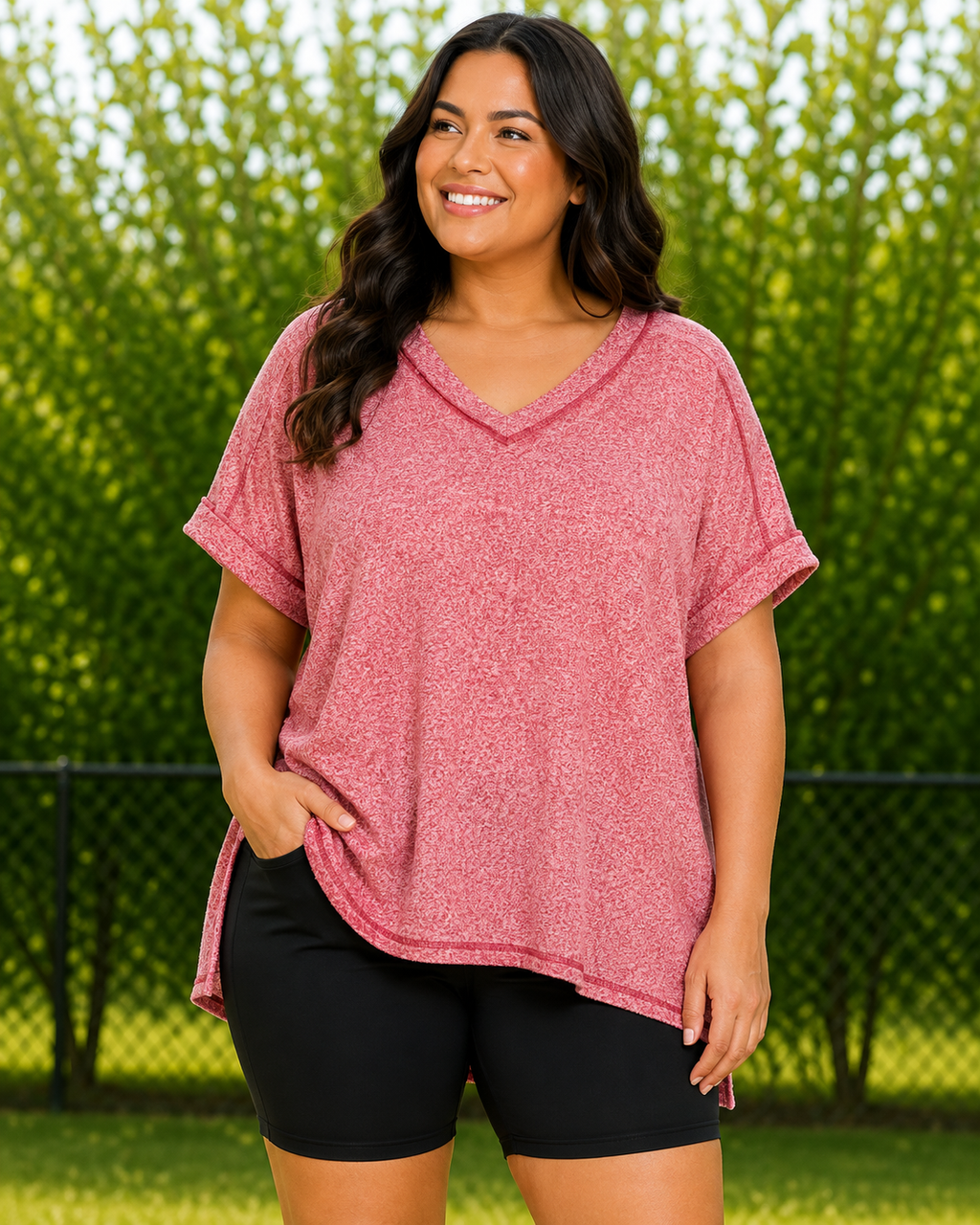 A woman wearing a heathered dusty rose short-sleeve V-neck top and black bike shorts, standing outdoors in front of a green hedge and chain-link fence.
