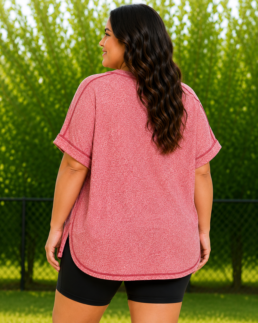 A woman viewed from the back wearing a heathered dusty rose short-sleeve top and black shorts, standing outdoors in front of a green hedge and chain-link fence.