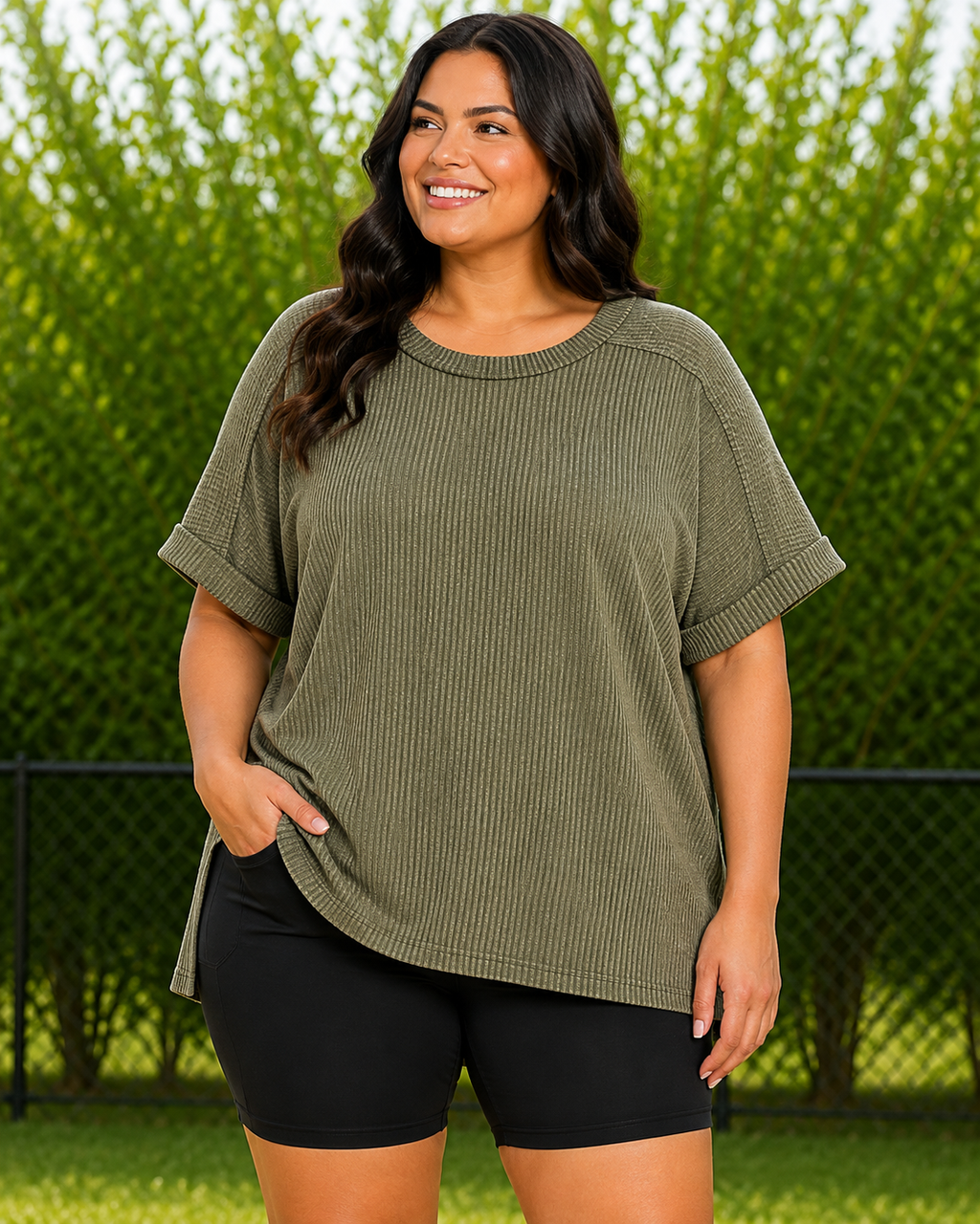 A woman wearing an olive green ribbed short-sleeve t-shirt and black bike shorts, standing outdoors in front of a green hedge and chain-link fence.